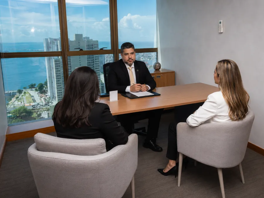 Dr. Capistrano Barros atendendo duas pessoas em sala com mesa, poltronas e janela envidraçada com vista para o mar do Recife