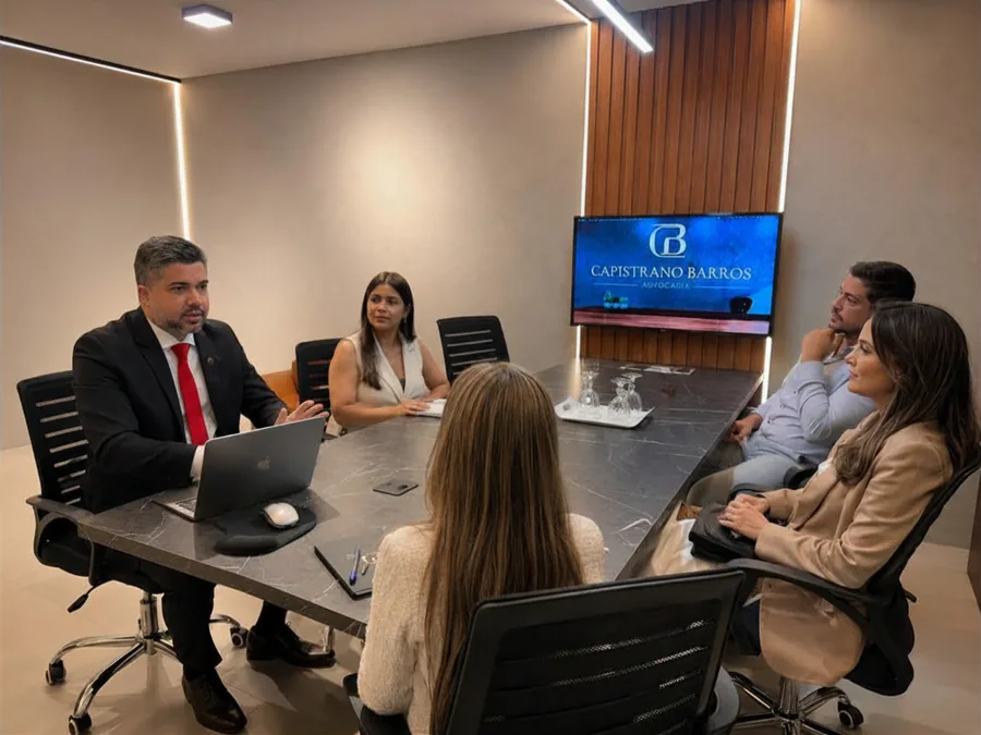 Reunião na sala de reuniões do escritório, com o advogado à mesa e a marca Capistrano Barros exibida na televisão
